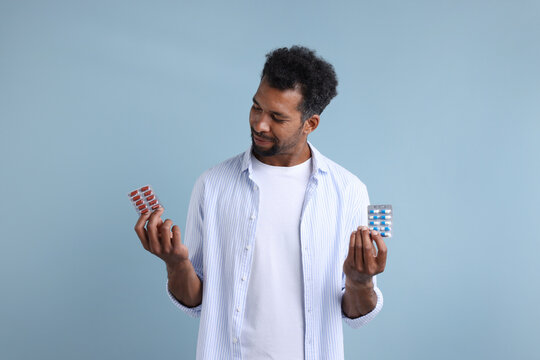 Man choosing between two blisters of pills on light blue background