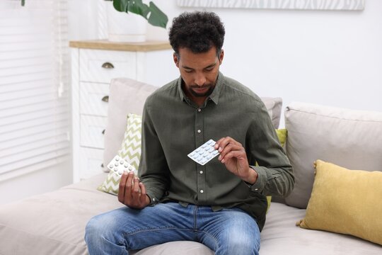 Man choosing between two blisters of pills on sofa at home