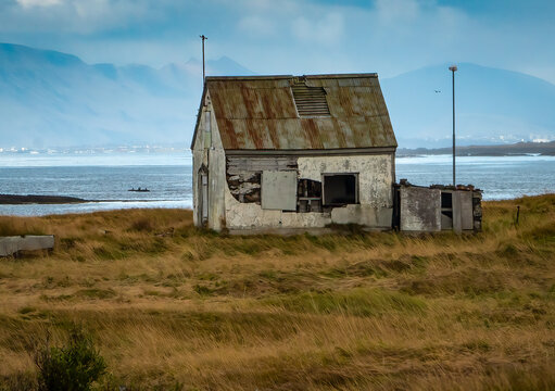 View of a weathered, dilapidated house with a rusted metal roof and peeling walls, situated in a grassy field overlooking the sea with distant mountains in Budir, Snaefellsbaer, Iceland.