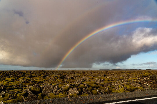 View of a vibrant double rainbow arching over a vast, moss-covered lava field under a dramatic cloudy sky in Grindavik, Grindavikurbaer, Iceland.