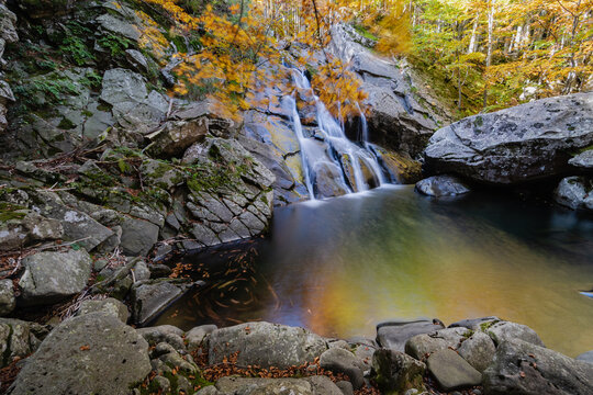 View of the Lagoni waterfall cascading into a rocky pool surrounded by vibrant autumn foliage and moss-covered stones in Corniglio, Emilia-Romagna, Italy.