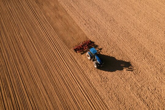 Blue tractor plows brown earth, creating straight furrows in a vast agricultural field