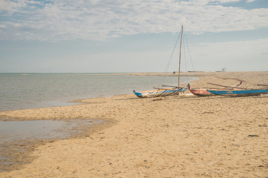 View of traditional wooden outrigger boats resting on the sandy shore of a calm beach under a soft cloudy sky in Morondava, Menabe, Madagascar.