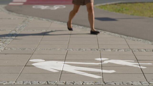 Pedestrian symbols painted on city sidewalk with woman walking in the background, marked pedestrian and bicycle paths separating urban mobility zones on sunny summer day