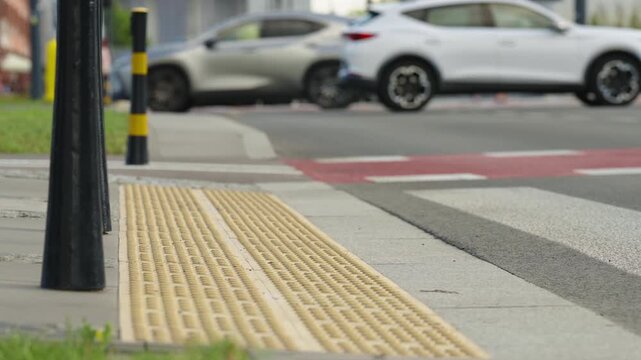 Close up of tactile paving at urban pedestrian crossing with cars moving in background, textured sidewalk surface, crosswalk markings and modern city street infrastructure