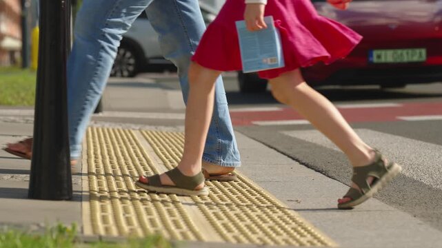 Close up of pedestrians crossing city street over tactile paving and marked crosswalk, multiple people walking through busy urban intersection with vehicle traffic on sunny summer day