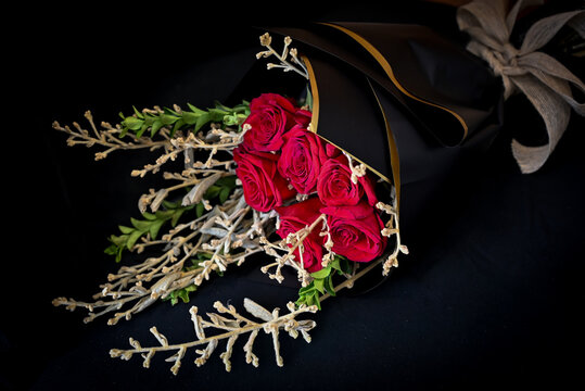 elegant red roses bouquet in black wrapping paper with white ribbon, romantic flower gift resting on dark chair, floral design close up