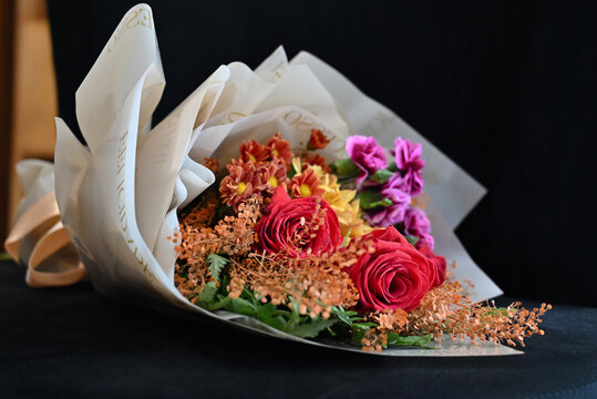 vibrant flower bouquet wrapped in white tissue paper, floral arrangement with red roses, purple carnations and yellow alstroemeria resting on dark surface, gift bouquet close up