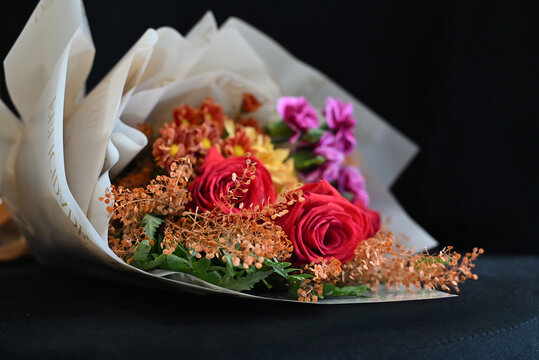 vibrant flower bouquet wrapped in white tissue paper, floral arrangement with red roses, purple carnations and yellow alstroemeria resting on dark surface, gift bouquet close up