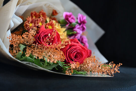 vibrant flower bouquet wrapped in white tissue paper, floral arrangement with red roses, purple carnations and yellow alstroemeria resting on dark surface, gift bouquet close up