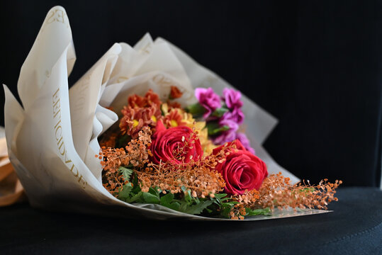 vibrant flower bouquet wrapped in white tissue paper, floral arrangement with red roses, purple carnations and yellow alstroemeria resting on dark surface, gift bouquet close up