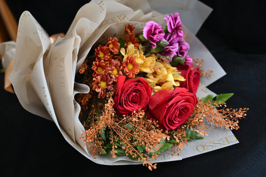 vibrant flower bouquet wrapped in white tissue paper, floral arrangement with red roses, purple carnations and yellow alstroemeria resting on dark surface, gift bouquet close up