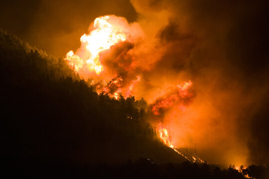 View of a forest fire burning on a hillside at night with intense orange flames and thick smoke rising above the dark silhouette of trees in Omis, Split-Dalmatia County, Croatia.