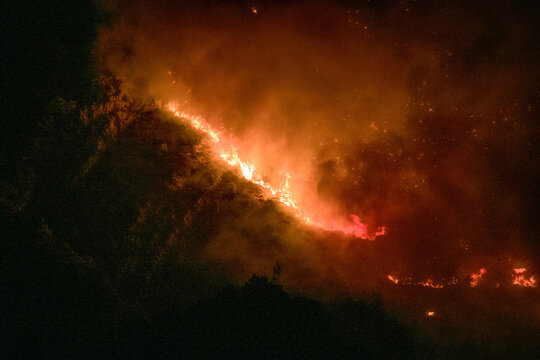 View of a wildfire burning on the hillside at night, with bright orange flames and thick smoke rising into the dark sky in Omis, Split-Dalmatia County, Croatia.