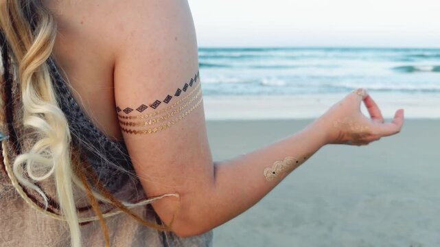 Woman performing a yoga mudra hand gesture with temporary golden flash tattoos on her arm and hand, focusing on mindfulness and inner peace during a meditation session on a sandy beach