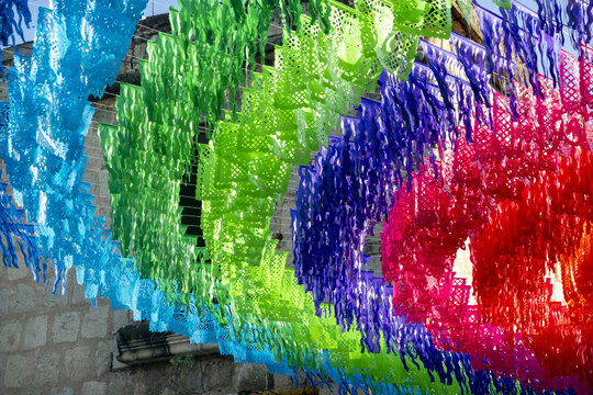 View of vibrant colorful Papel Picado decorations hanging in a street against a historic stone wall in Oaxaca, Oaxaca, Mexico.