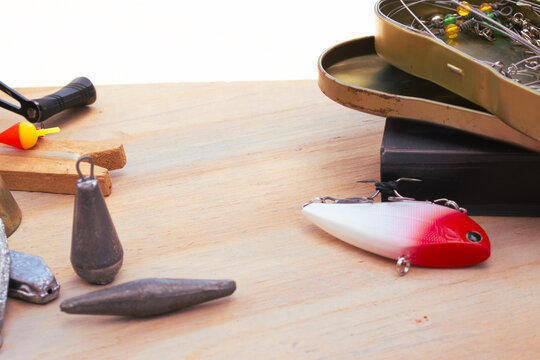 Fishing gear laid out on a wooden background: red-and-white lure, lead weights, bobber and open tackle box with hooks, swivels and pliers for an angling setup