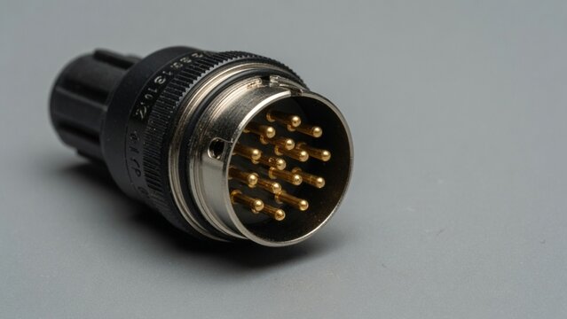 Black multi-pin circular serial connector with visible metal contact pins resting on a clean light grey surface, industrial electronic component, vintage audio plug close-up