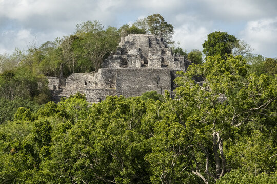 View of the ancient Mayan pyramid of Palenque rising above the dense green jungle canopy under a partly cloudy sky in Palenque, Chiapas, Mexico.