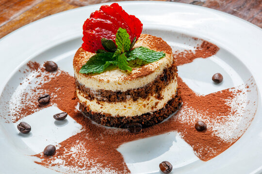Gourmet layered coffee and chocolate dessert topped with fresh strawberry and mint, served on a white plate with cocoa powder and coffee beans.