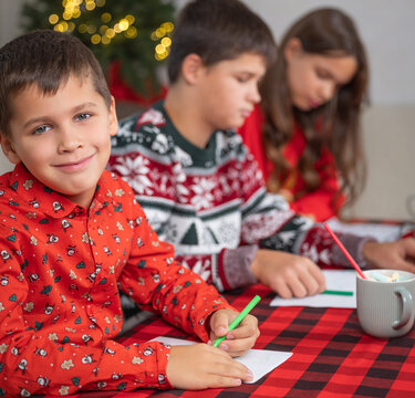 Close view of three children in holiday pajamas gathered at red and black checkered table, with youngest boy smiling at camera while siblings focus on drawing and coloring, background shows blurred