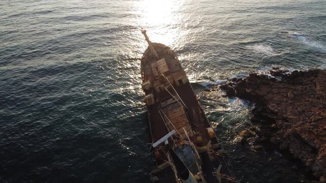 Aerial view of rusted shipwreck partially submerged near rocky coastline with clear turquoise water and waves crashing against shore. Aerial view of coastal wreckage.