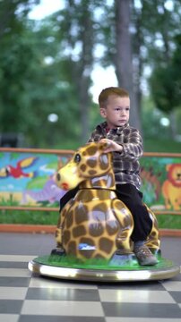 Young boy rides a giraffe-shaped kiddie car at an outdoor amusement park, holding the handles.