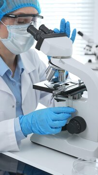 Female scientist analyzing sample through microscope, wearing protective lab gear during medical research investigation. Medicine, laboratory and science concept