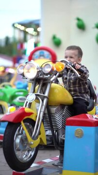 Happy little child holds handlebars of a bright carnival motorcycle ride at dusk, colorful park attractions and glowing lights behind him.