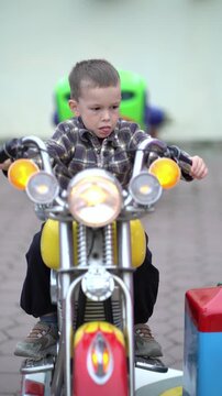 Cute smiling boy rides a colorful yellow motorcycle attraction at an amusement park with lights and other kiddie rides in the background.