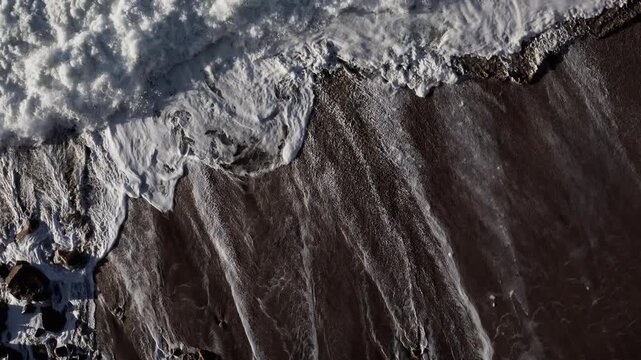 Aerial zoom in on abstract texture of foamy Atlantic Ocean waves washing onto sandy coast by Farol da Nazare in Portugal, beautiful natural background pattern of white sea foam, 4K aerial drone shot