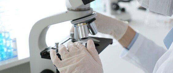 Gloved hands adjusting a microscope in a bright laboratory, emphasizing precision and focus in...
