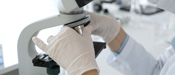 White gloved hands of scientist professional adjusting a microscope in a bright laboratory,...