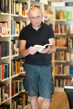 Focused middle-aged guy browsing through literature volumes on shelves in cozy bookshop