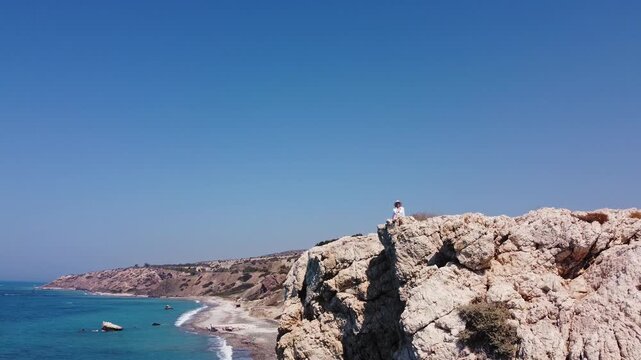 Person sits atop rugged coastal cliffs overlooking turquoise sea and pebble beach with distant hills under clear blue sky. Woman seated above a pebble beach.