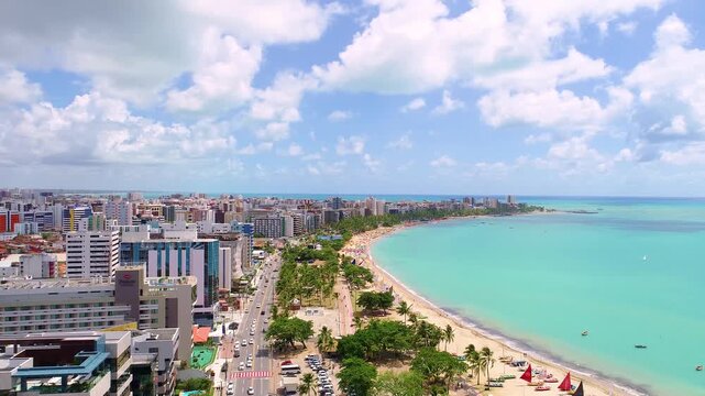 4K aerial view of Maceio, Alagoas, Brazil, featuring turquoise Atlantic waters, tropical beachfront, palm trees, avenue and modern buildings along the coast.