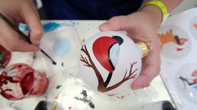 A video of a woman sitting at a craft table painting a Christmas tree ornament depicting a bullfinch.