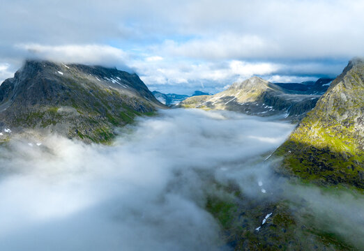 Aerial view of Trollstigen mountain pass surrounded by dramatic peaks, lush green slopes, and low-hanging clouds in Andalsnes, More og Romsdal, Norway.