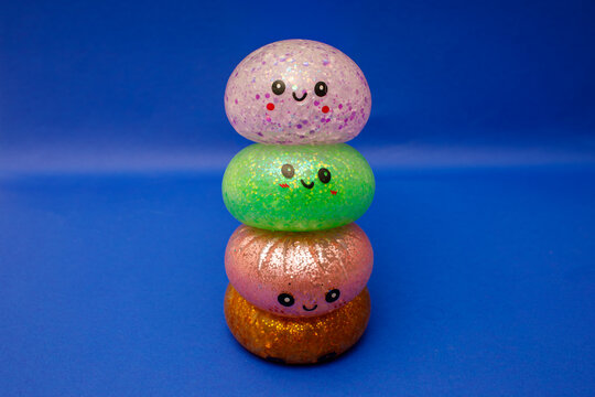 Stacks of glittery dumpling toys with kawaii smiling faces are arranged vertically against a deep blue background, featuring pink, green, coral, and bronze tones