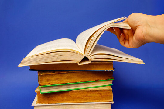 A hand flips the pages of an open book resting on a stack of closed hardcover books for a back to school concept, against a vibrant blue background
