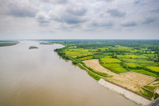 Aerial view of the Ganga Expressway construction site along the Ganges river, featuring lush green agricultural fields and a cloudy sky in Daharpur Kalan, Uttar Pradesh, India.