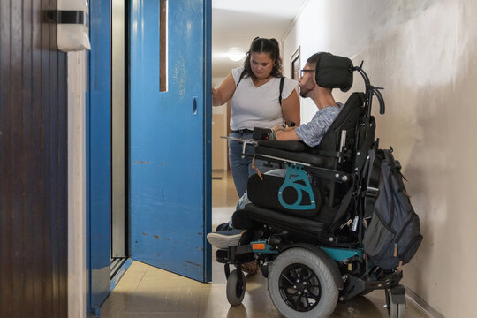 Powered wheelchair user entering an elevator independently, with a Personal assistant holding the door open in an accessible building.