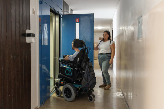 Person with disability entering an elevator with a Personal assistant holding the door open in apartment building.