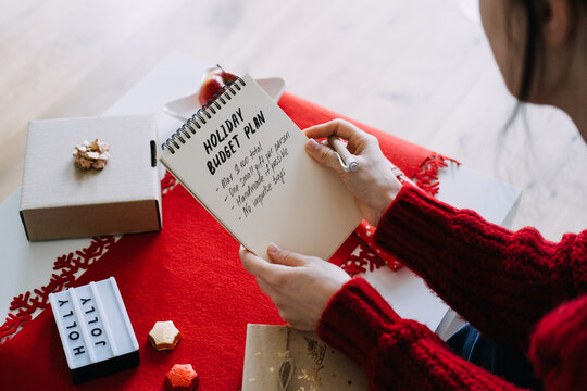 Hands hold notebook with HOLIDAY BUDGET PLAN above table with HOLLY JOLLY sign and wrapped gifts. Christmas no impulse buying, mindful christmas spending, budget conscious holiday shopping