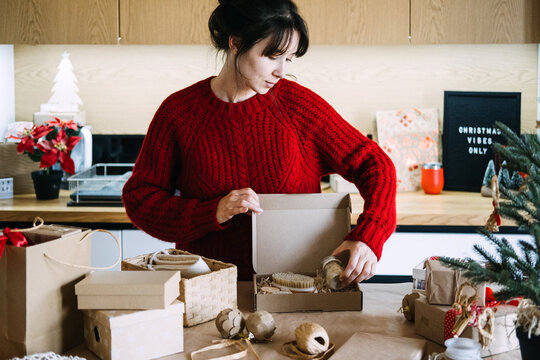 Woman places jar into open kraft box with brush in kitchen near CHRISTMAS VIBES ONLY sign. Checking christmas gift set before shipping, spa gift box quality check, verifying gift box contents