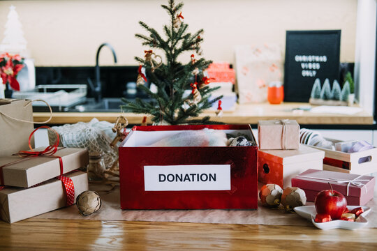 DONATION box filled with folded knitwear stands among wrapped presents near Christmas tree. Christmas charity donation, holiday volunteering concept, giving back to community christmas