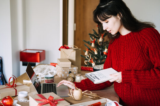 Woman holds notebook with CHRISTMAS SHOPPING LIST while using laptop among wrapped gifts. Christmas online shopping laptop, checking christmas list laptop, online gift shopping holiday