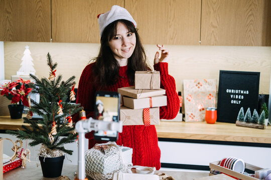 Phone on tripod records woman in Santa hat carrying stack of wrapped parcels in kitchen. Influencer marketing holiday season, small business video promotion, content creator business marketing