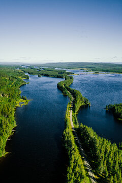 Aerial view of a winding road on a narrow strip of land separating two deep blue lakes surrounded by dense coniferous forests under a clear blue sky in Jamtland, Sweden.