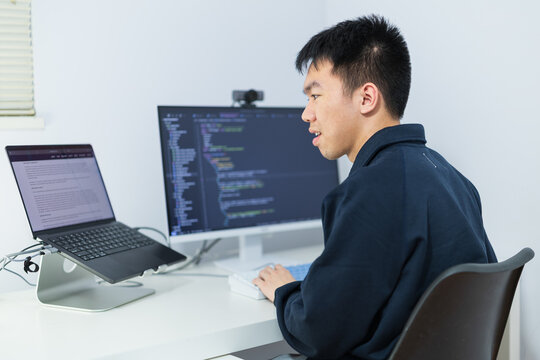 A young Asian man works remotely at a home desk while coding across a laptop and desktop monitor, for software development, technology careers, or work-from-home concepts.